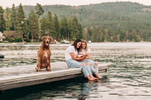 Family and their brown dog sitting together on wooden dock by mountain lake.