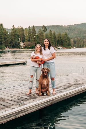Family portrait on lakeside dock with brown dog against mountain backdrop.