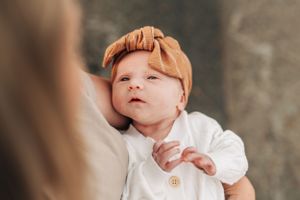 Baby wearing white outfit and orange headband looking up with curious expression.