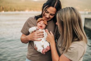 Two people sharing an intimate moment with a crying newborn by a waterfront at sunset.