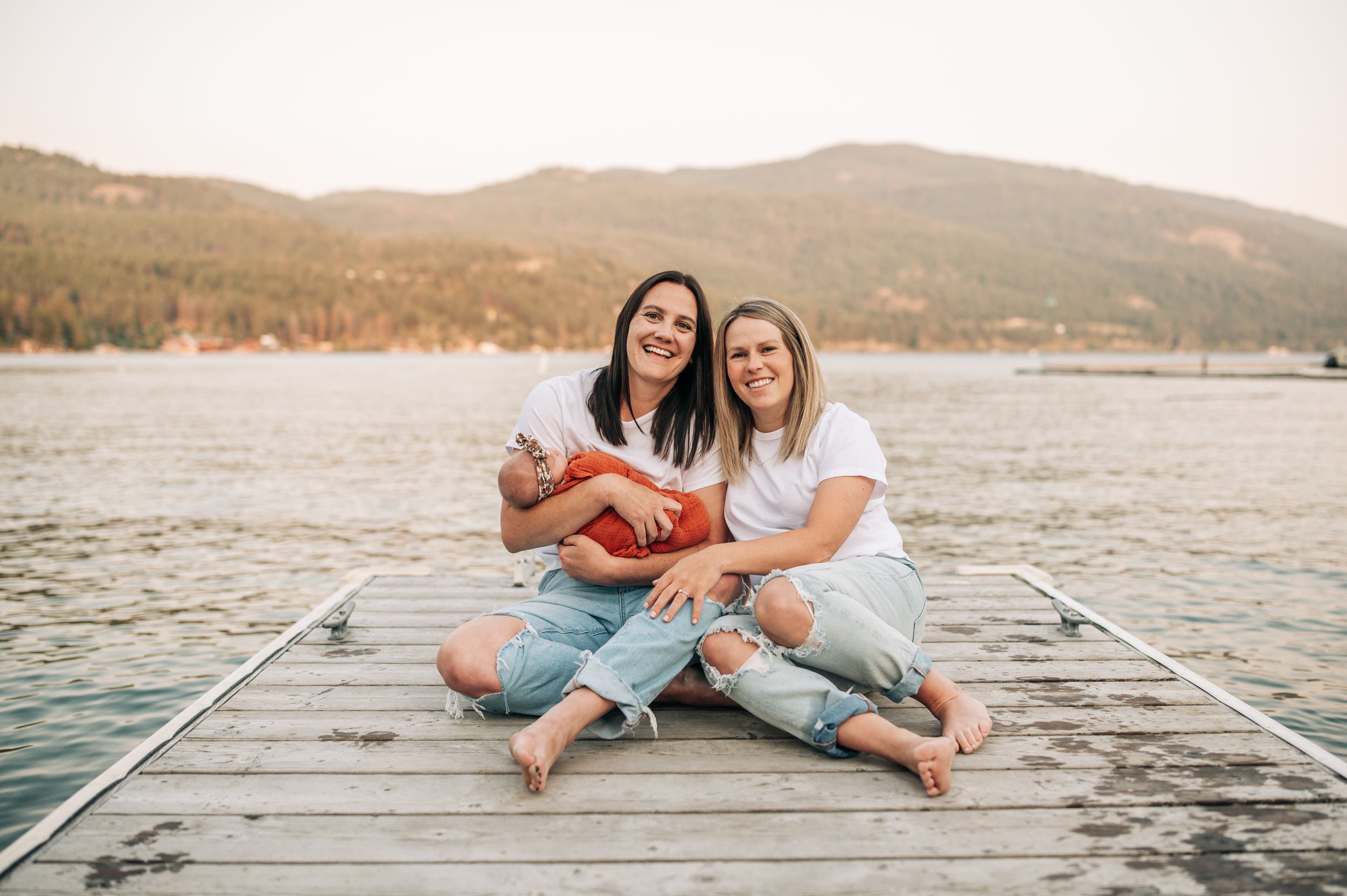 Two people sitting together on wooden dock by lake with mountains in background.