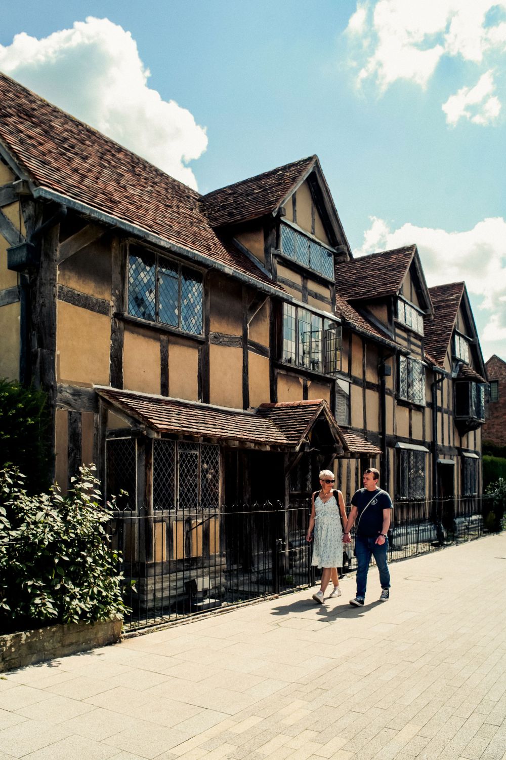 Couple walking past historic Tudor-style buildings with timber framing and leaded windows in a quaint town street.