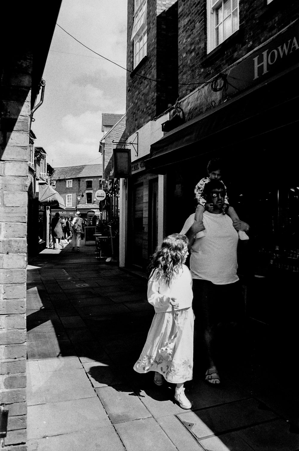 Black and white photo of people walking through a narrow shopping alley with brick buildings.
