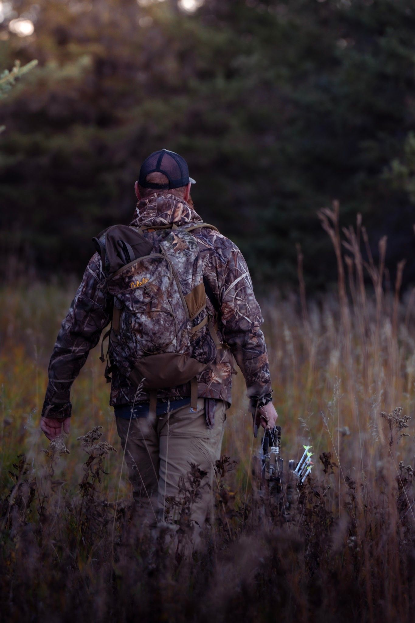 Hunter in camouflage gear walking through tall grass at dusk carrying equipment.