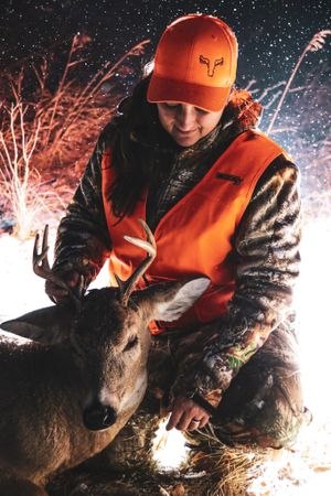 A hunter in orange safety vest examines a harvested deer in snowy nighttime conditions.