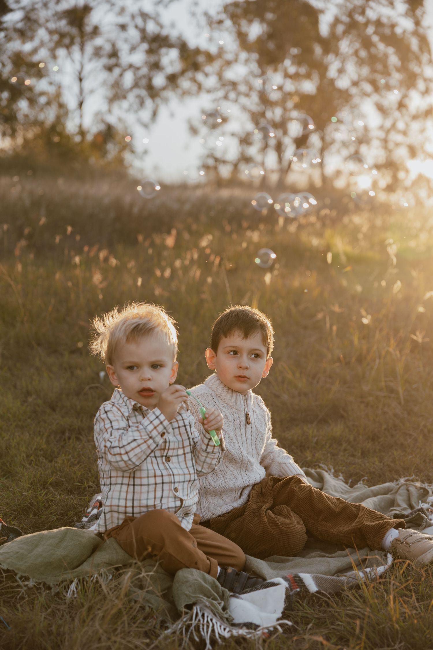 Two young boys sit on a picnic blanket in a golden sunset field while soap bubbles float around them.