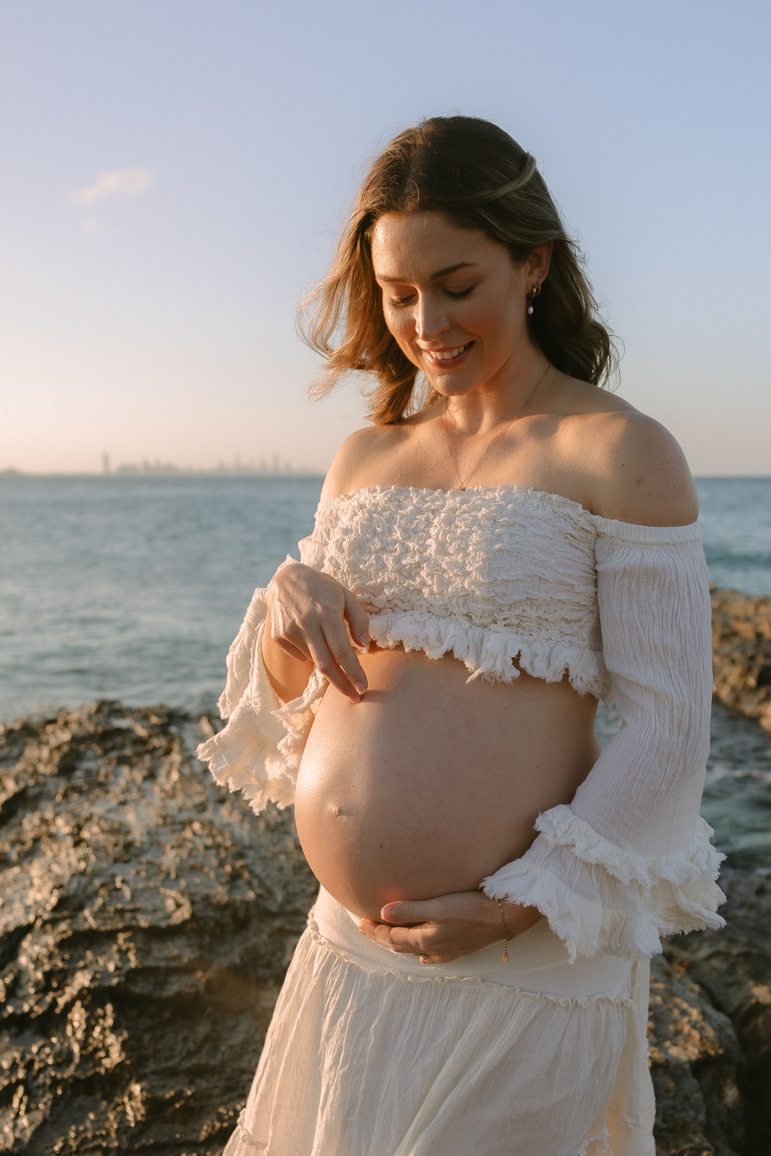 Smiling pregnant woman in a white off-shoulder top and skirt cradles her bare baby bump on rocks beside the ocean at sunset.