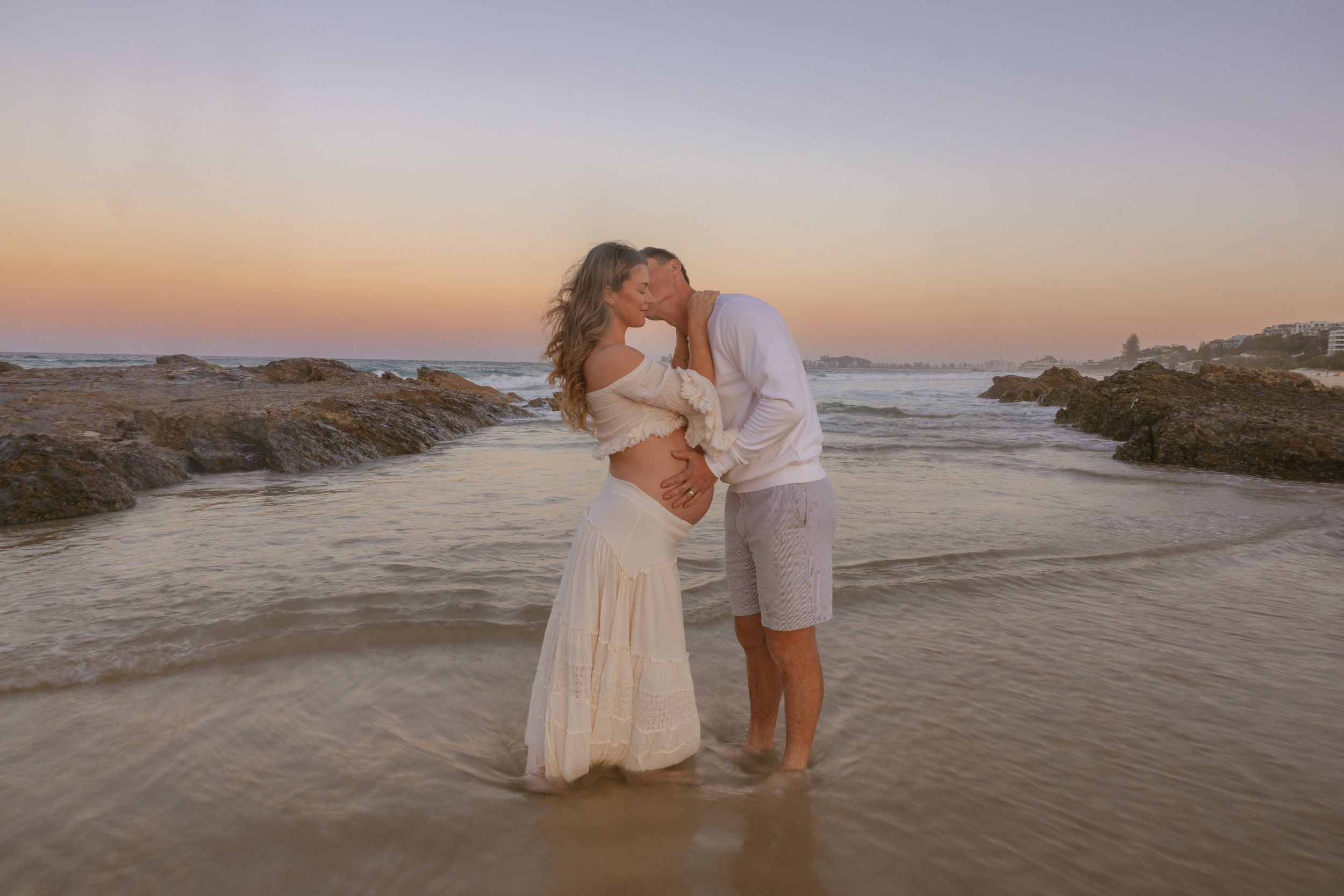 Pregnant woman and her partner share a kiss in shallow ocean water at sunset during a beach maternity photoshoot.