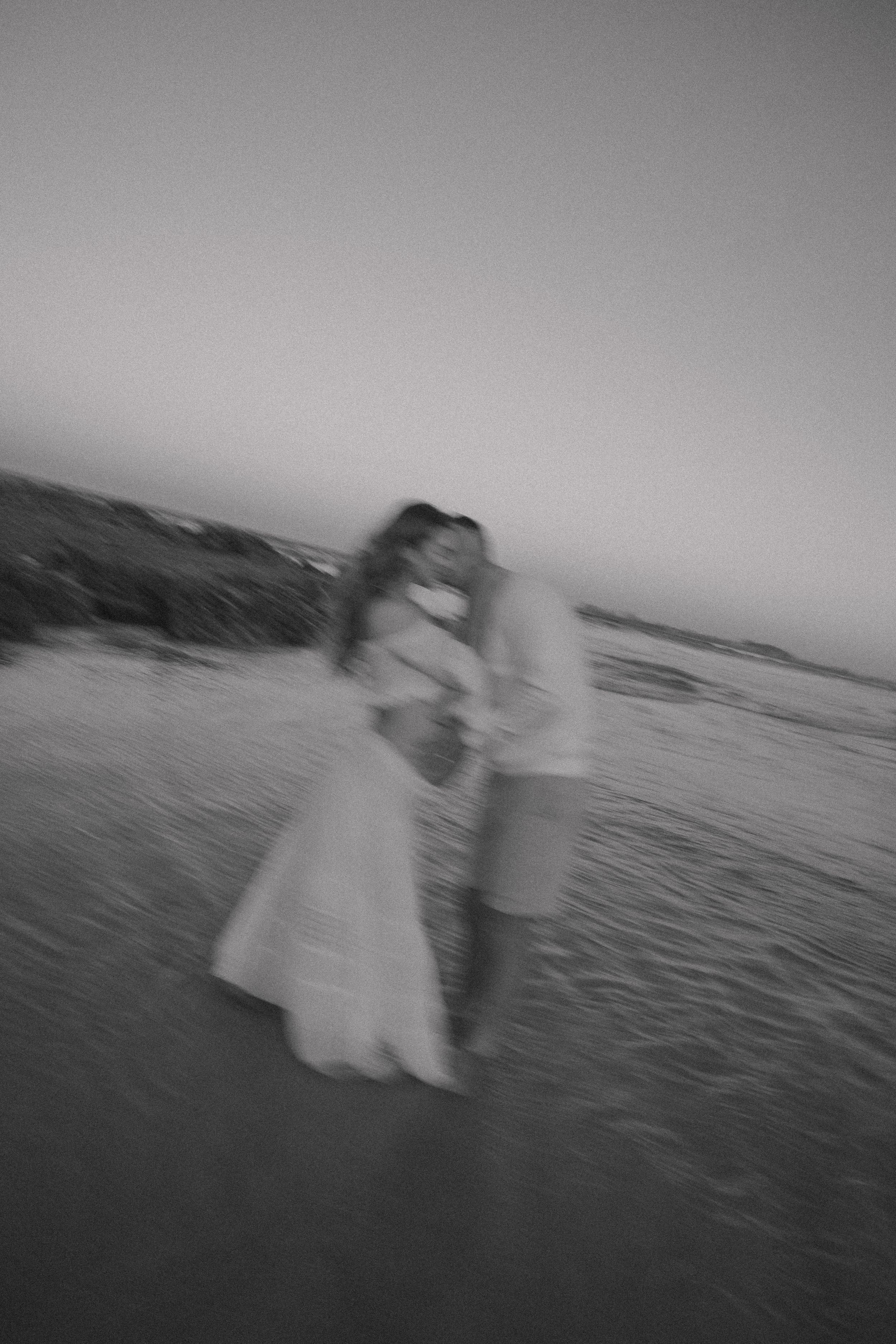 Blurry black and white beach maternity photo of an expecting couple embracing in the shallow waves at dusk.