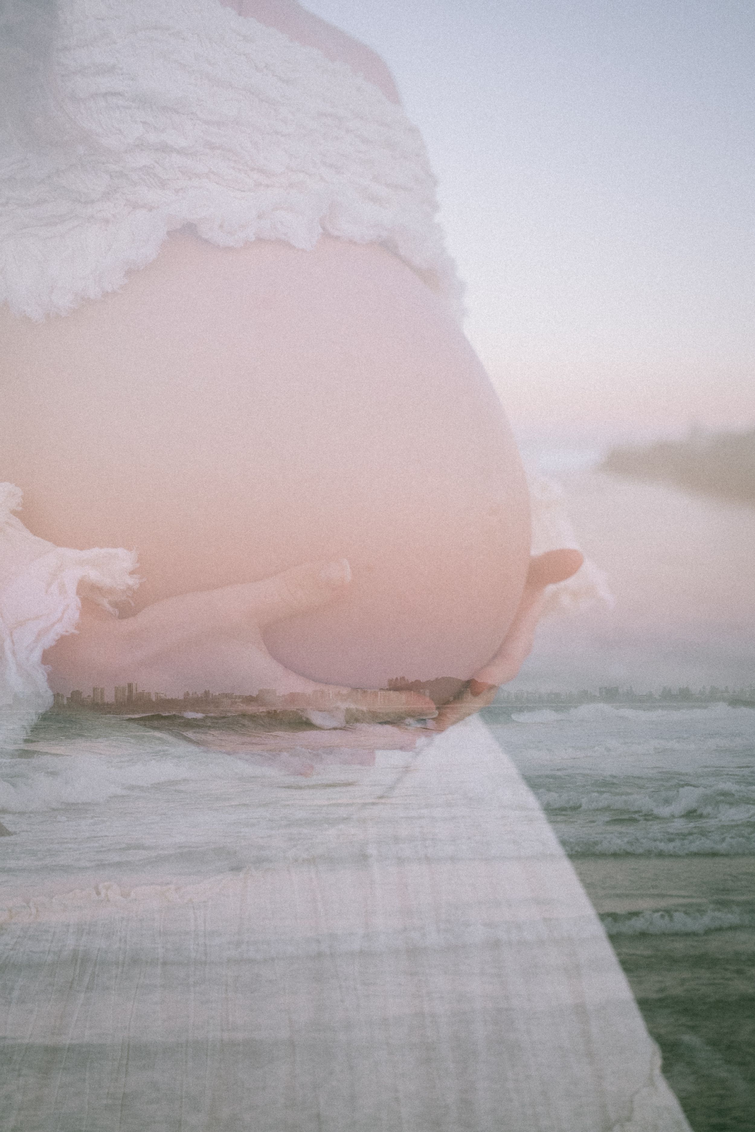 A double exposure maternity photo of a pregnant belly in a white lace top overlaid with a soft pastel ocean and pier scene.
