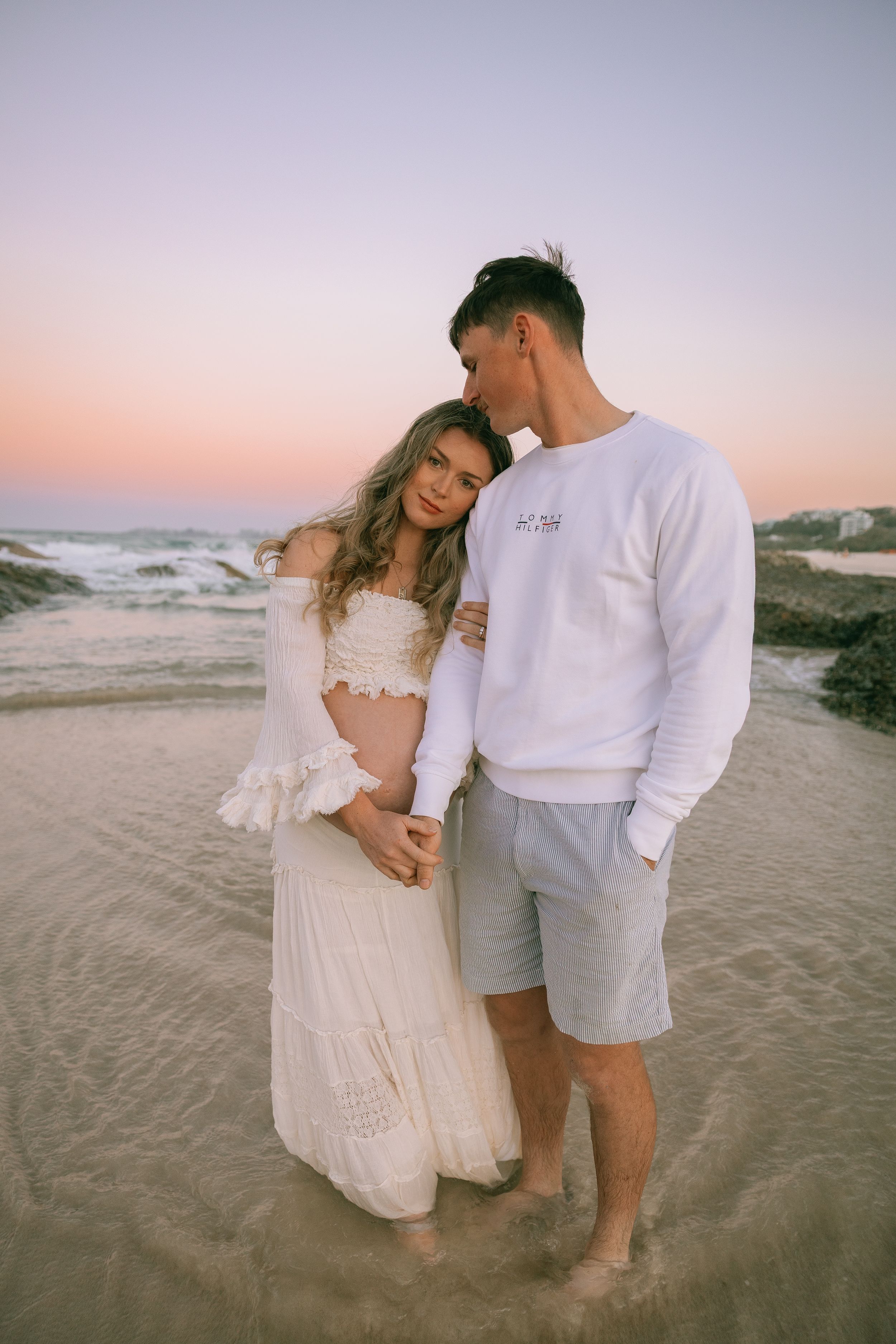 Pregnant woman in a cream boho outfit rests her head on her partner's shoulder on the beach at a pink sunset.