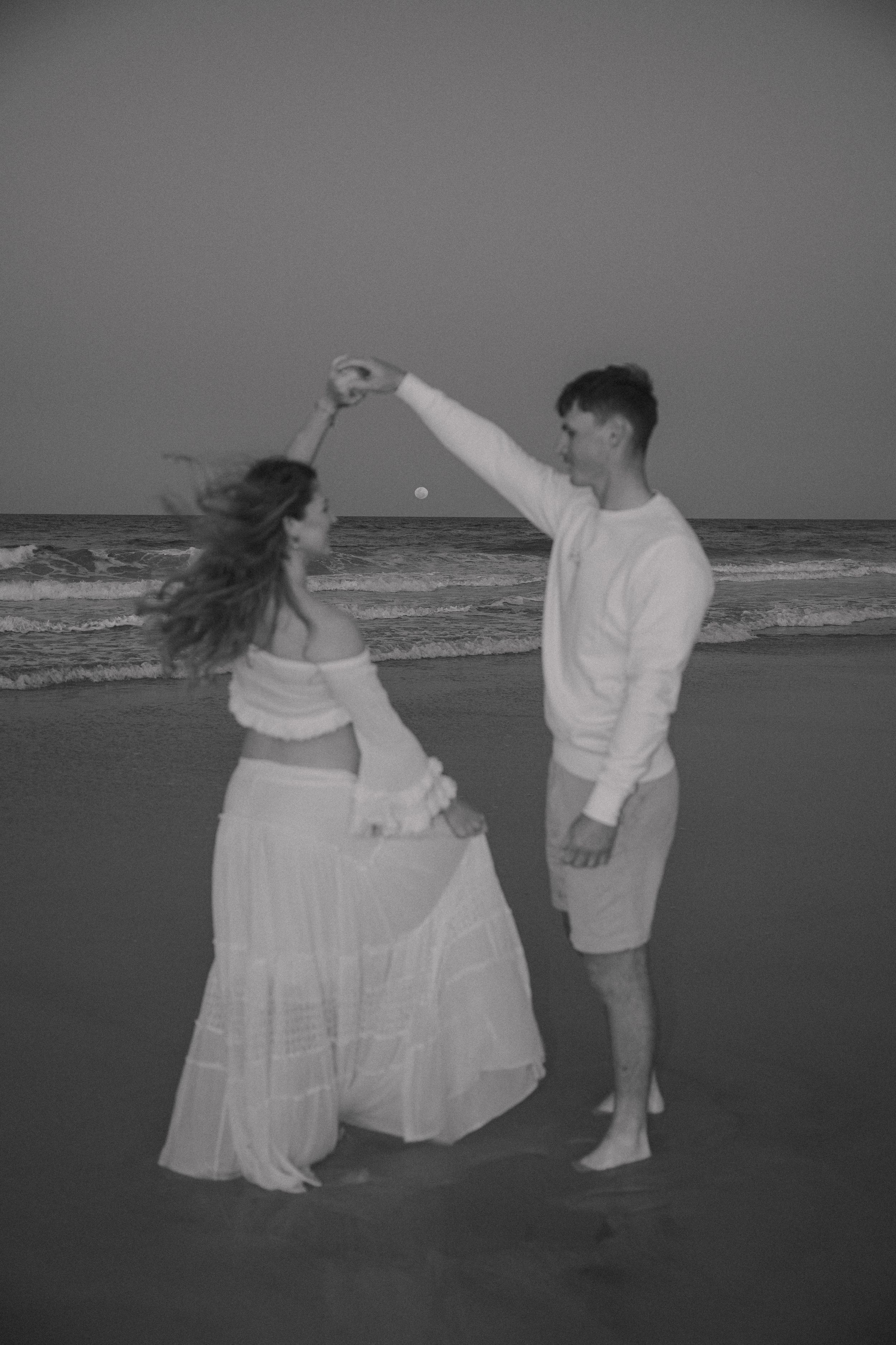 A black and white image of a couple dancing on the beach, the woman twirling in a flowing skirt as the moon rises over the sea.