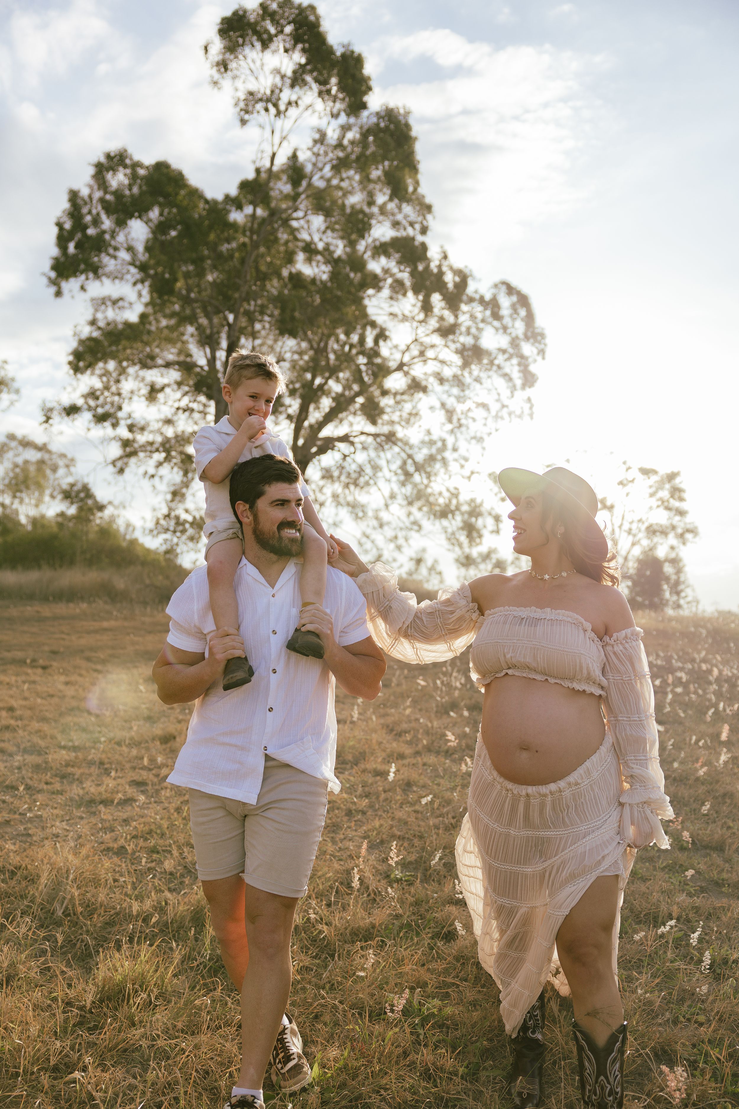 A smiling family walks through a golden field at sunset, with a boy on his dad's shoulders and a pregnant mother in a white outfit.