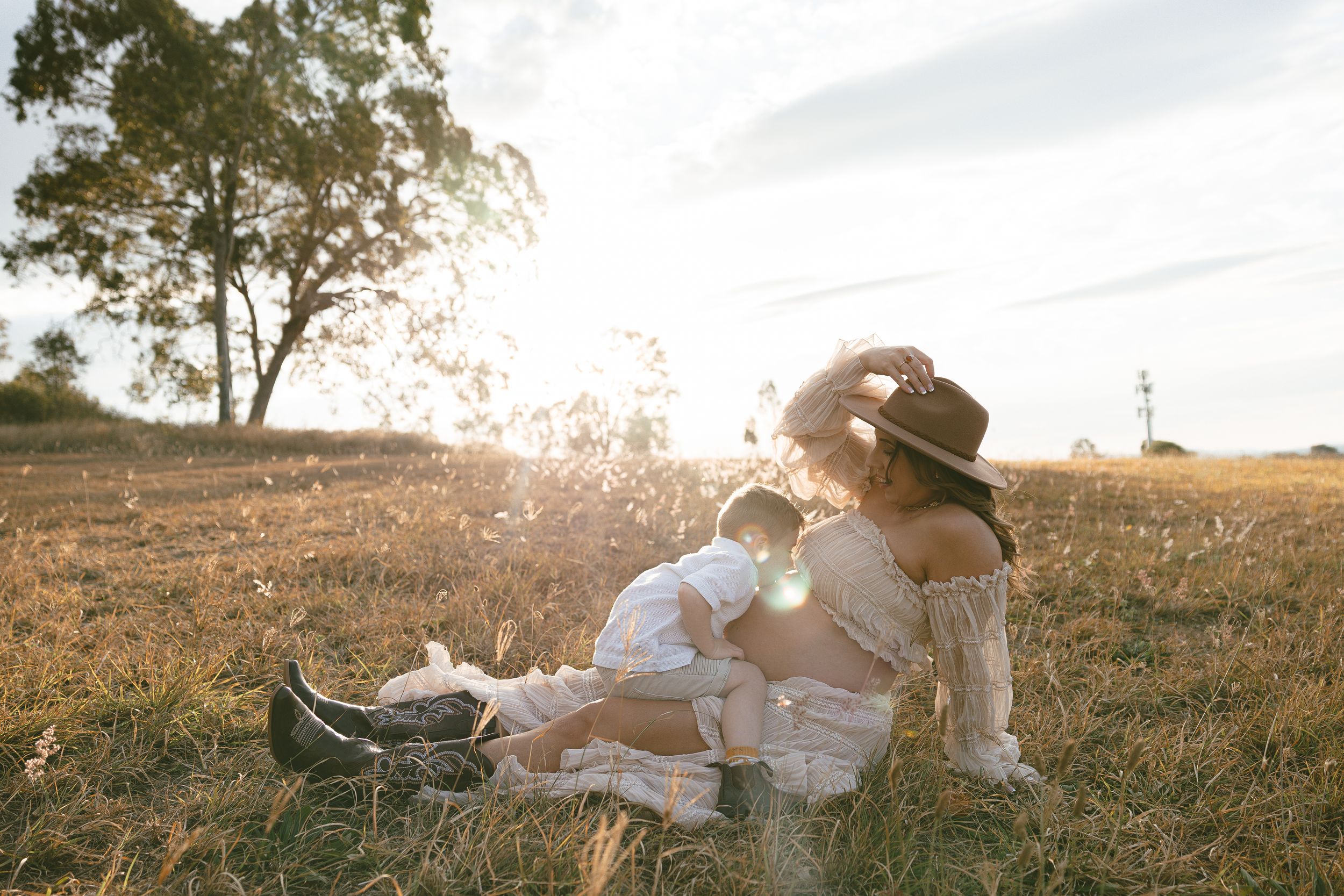 A pregnant mother in a hat and cowboy boots sits in a sunlit grassy field cuddling her toddler during a golden hour maternity session.