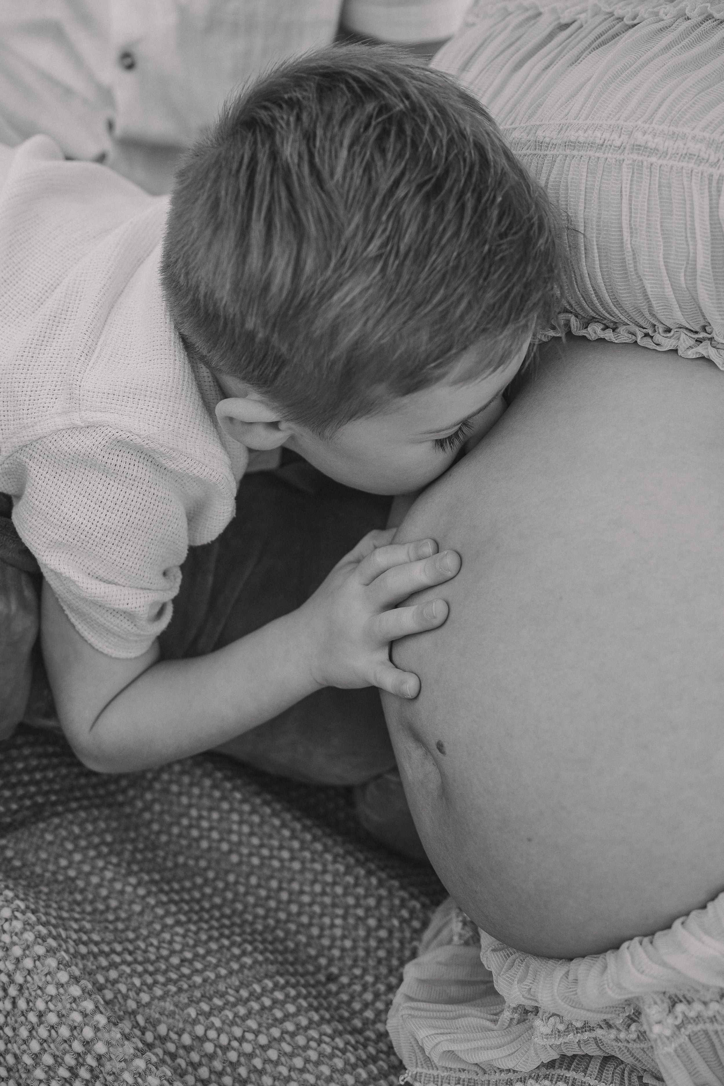 Black and white photo of a young boy leaning in to kiss his mother's pregnant belly during a maternity photoshoot.