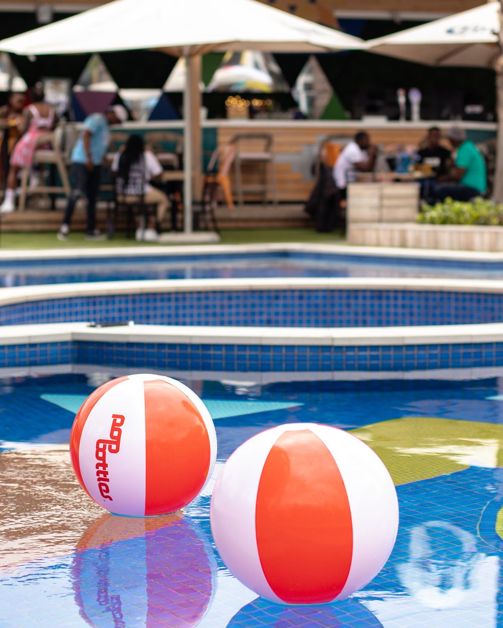 Colorful red and white beach balls floating in a blue swimming pool with umbrellas in the background.