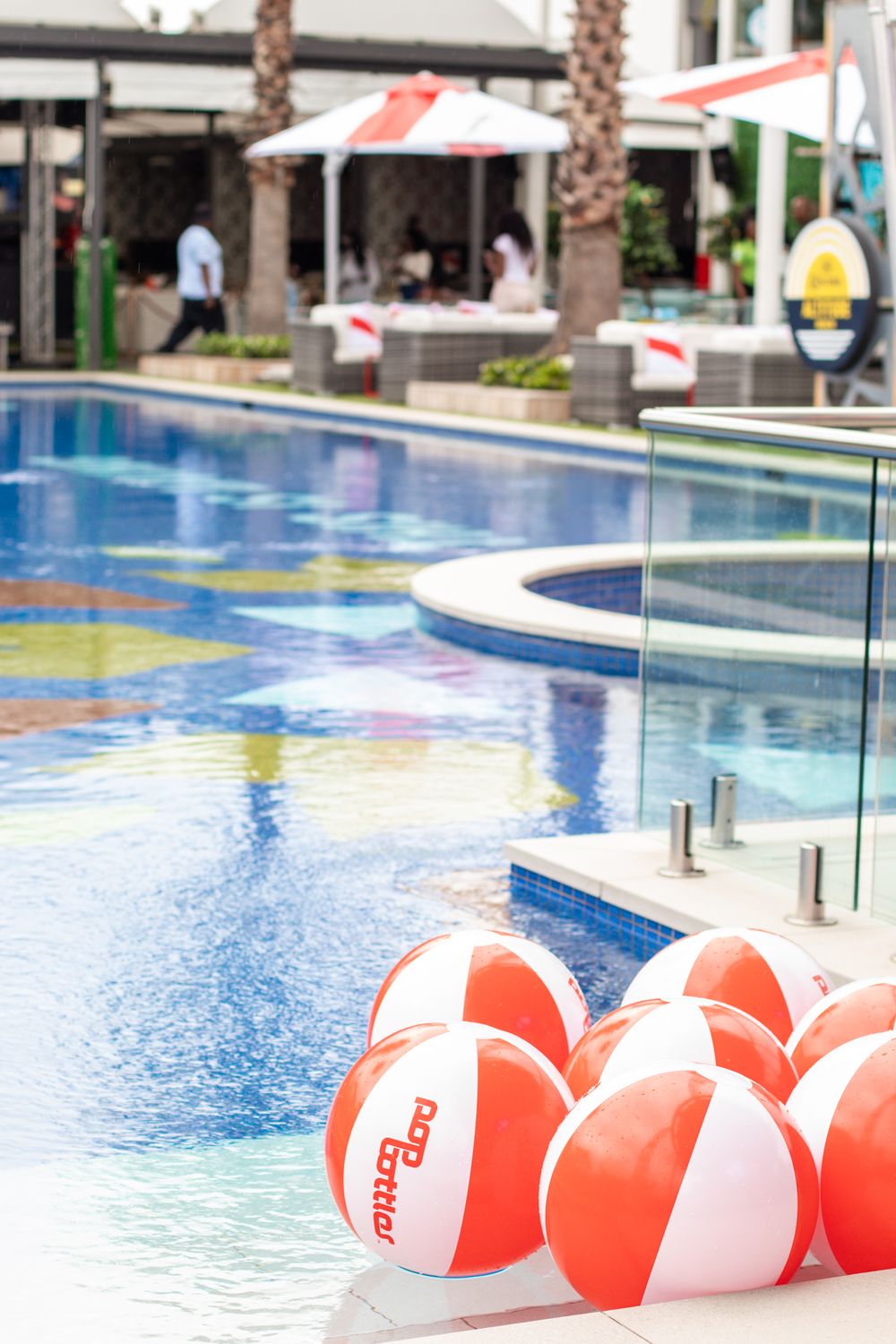 Red and white striped beach balls floating in a resort swimming pool with palm trees.