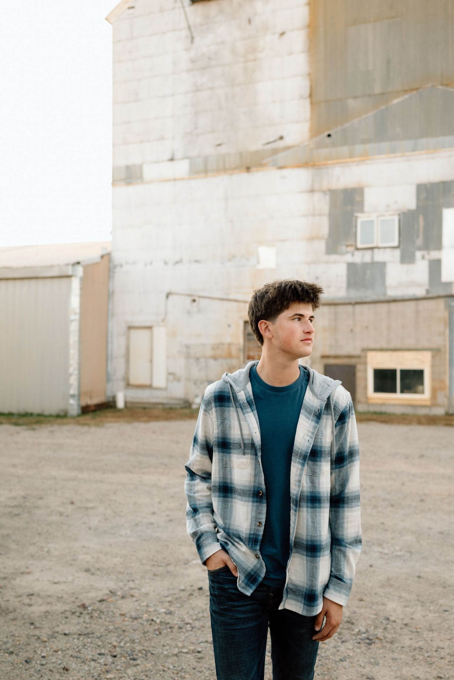 Someone in a plaid shirt and jeans poses against an industrial building exterior.