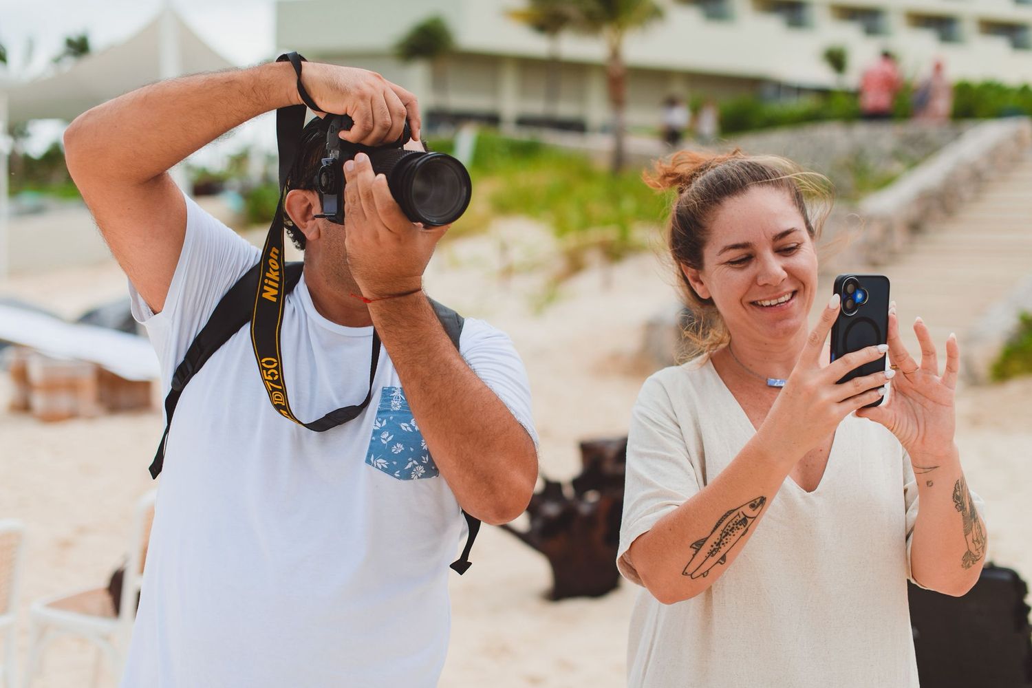 Man photographing with DSLR camera while woman takes photo on smartphone at a beach resort.