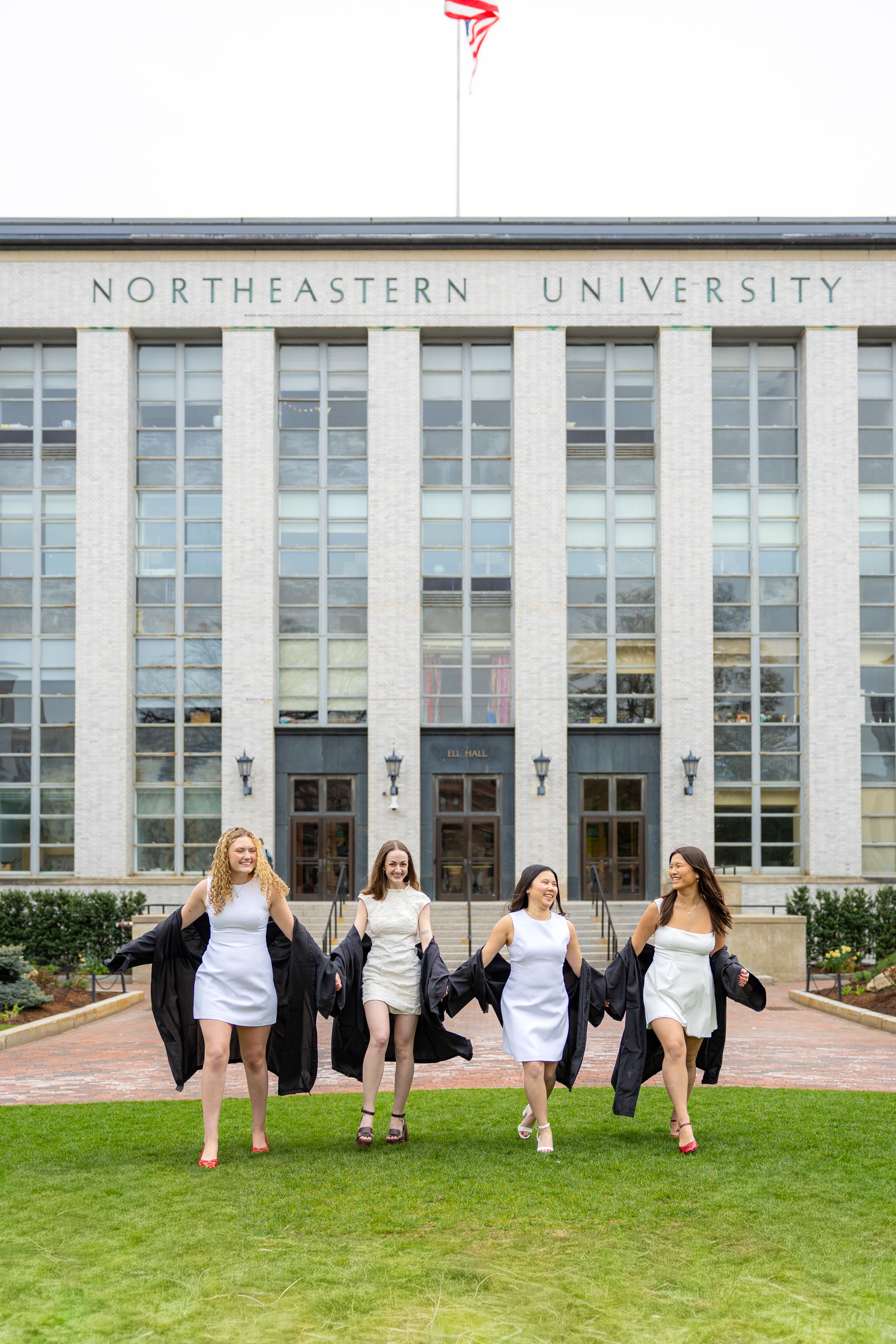 Four women in white dresses holding graduation gowns walk joyfully in front of Northeastern University building.