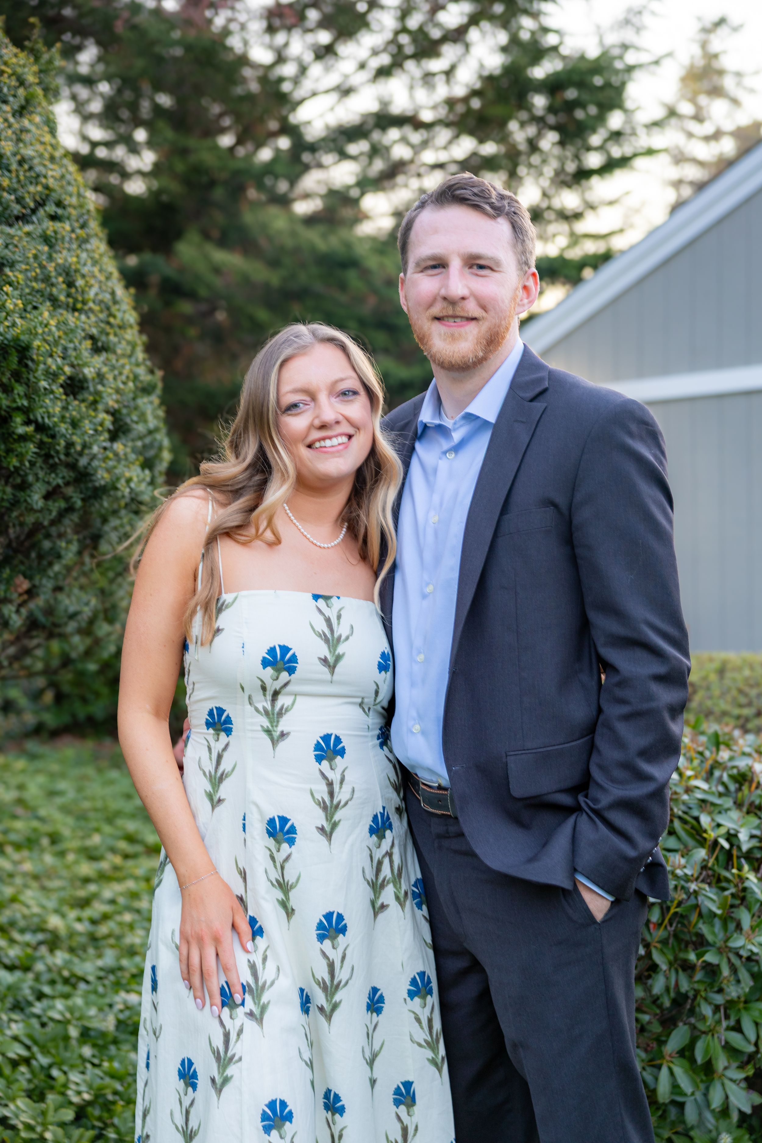 Smiling couple posing outdoors, woman in floral sundress and man in navy suit, with green hedges behind them.