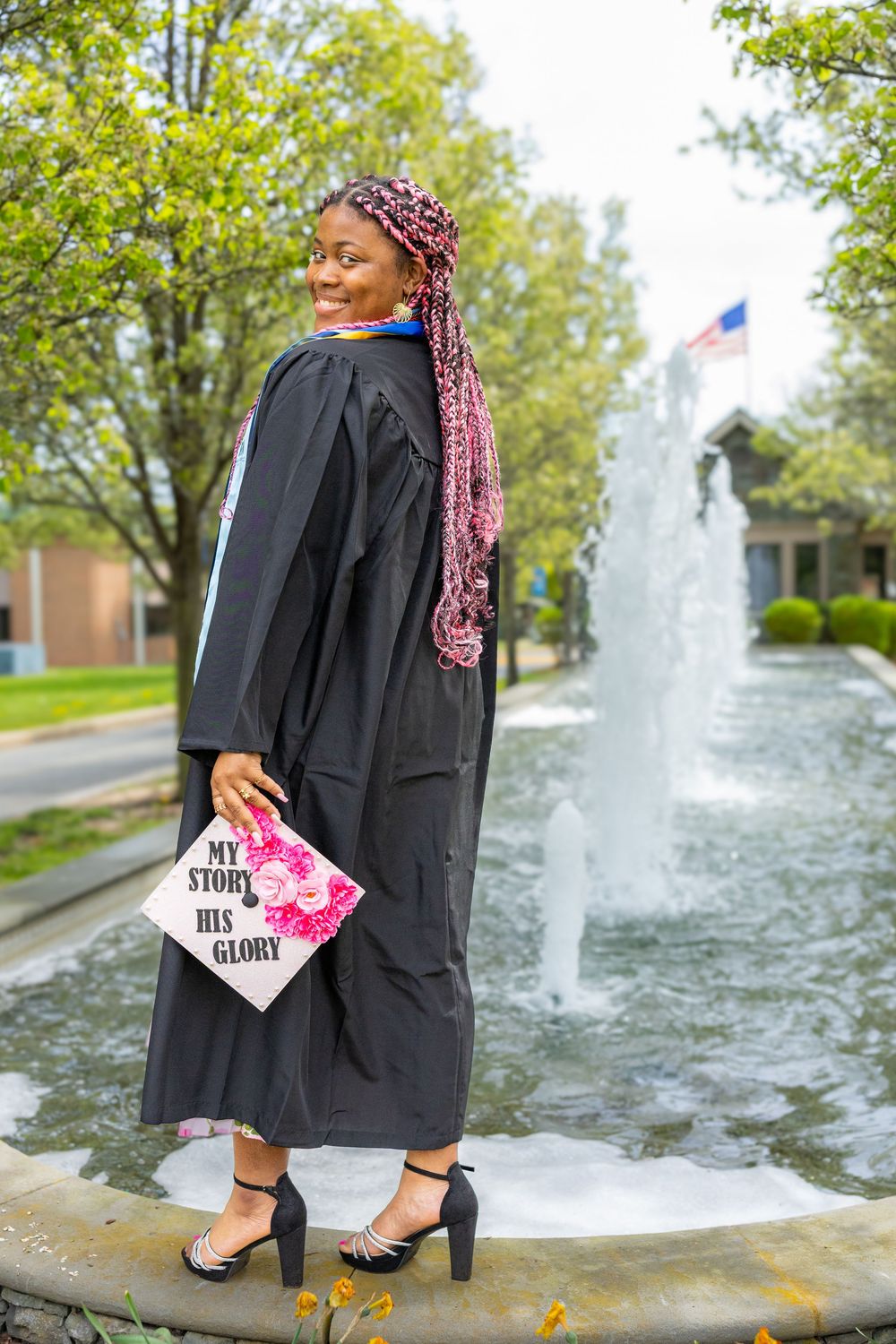 Graduate in black cap and gown poses with decorated graduation cap by fountain on sunny campus day.