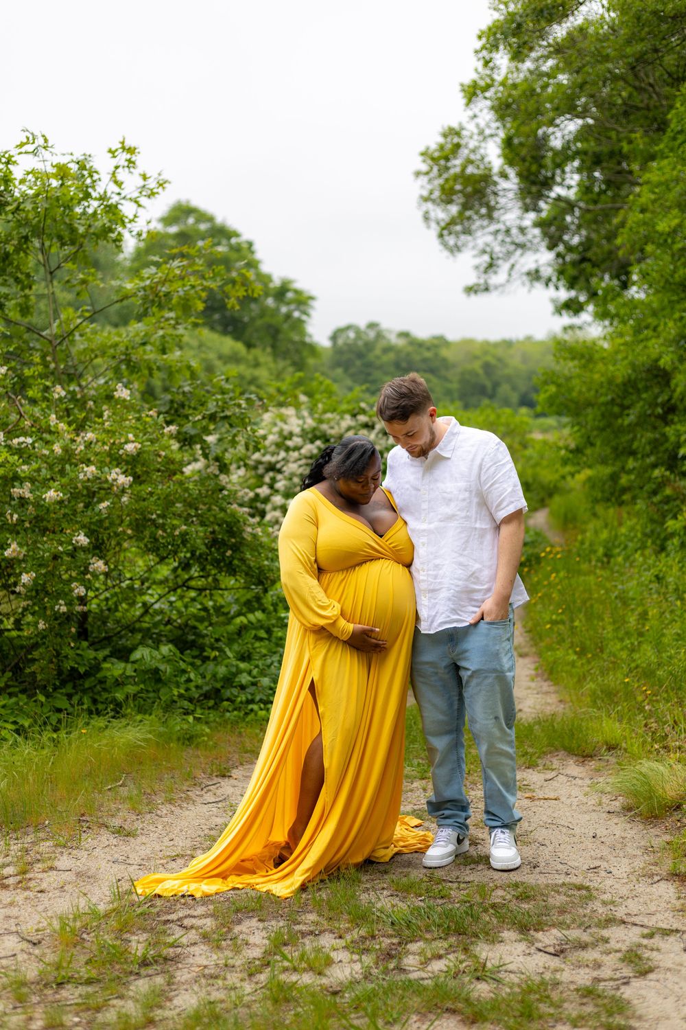 A couple shares an intimate moment on a dirt path surrounded by lush greenery while wearing a flowing yellow dress and white shirt.