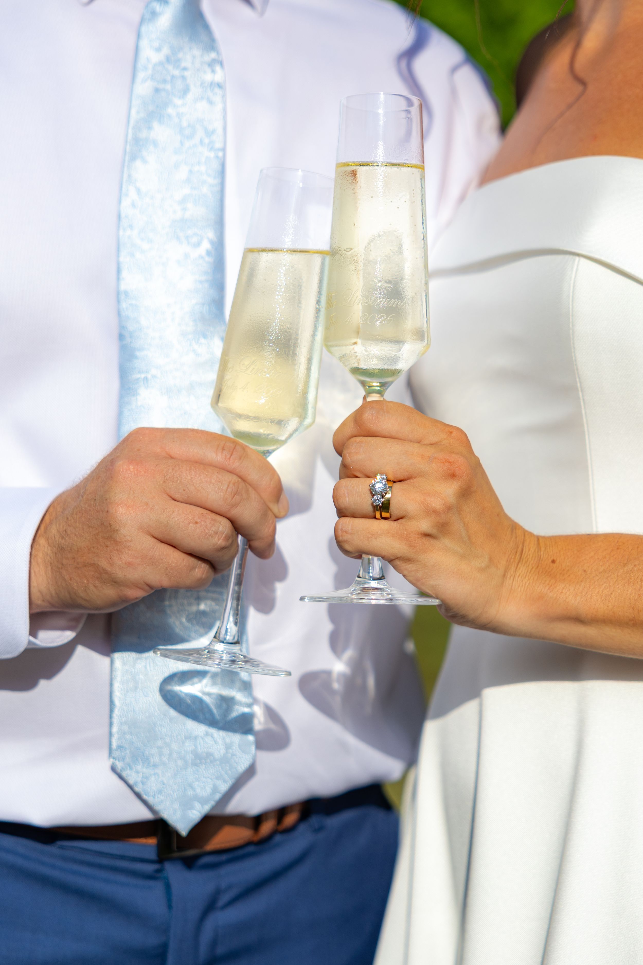 Newlywed couple toasting with champagne flutes, bride wearing diamond ring in white dress, groom in blue tie.