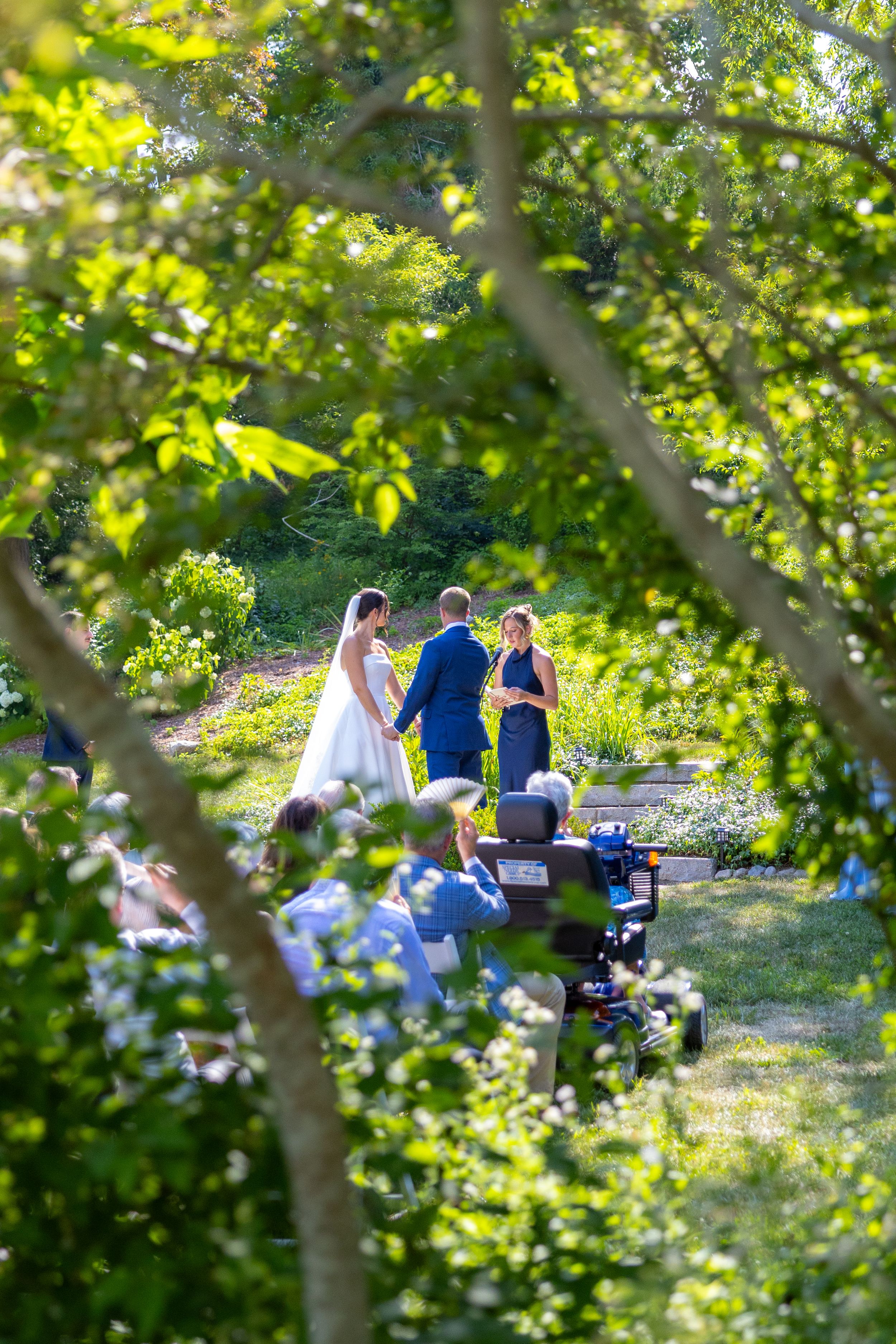 Outdoor garden wedding ceremony with bride and groom standing before officiant, framed by lush green trees and seated guests.