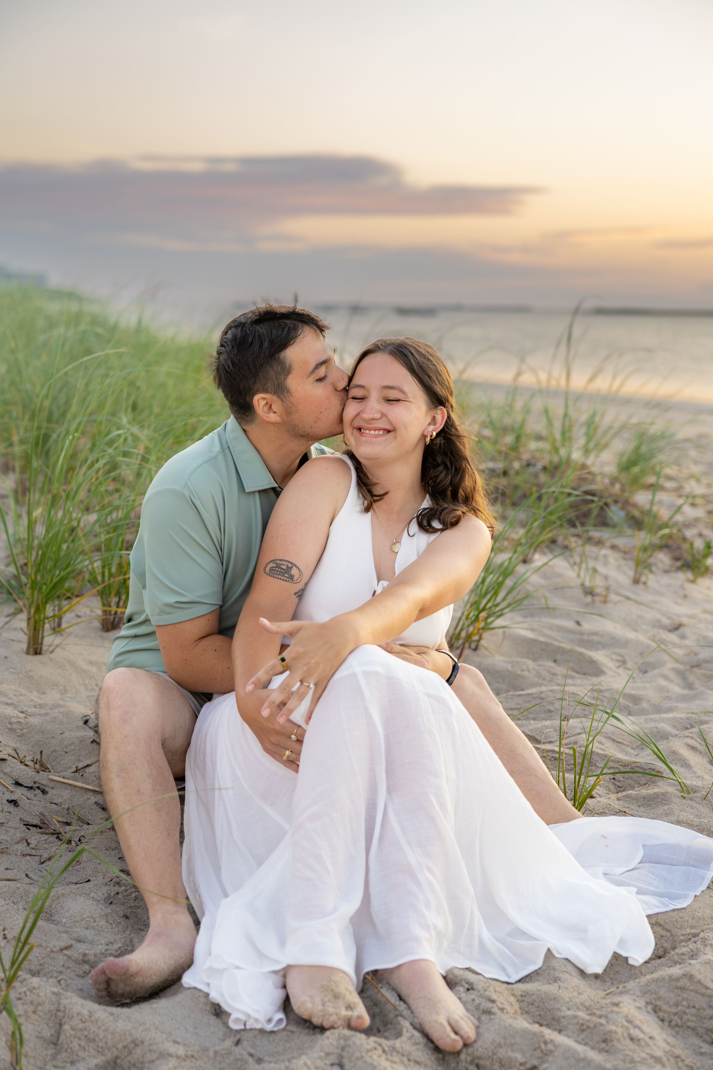 Couple sitting on beach sand dunes at sunset, man kissing woman's cheek as she smiles in white dress.