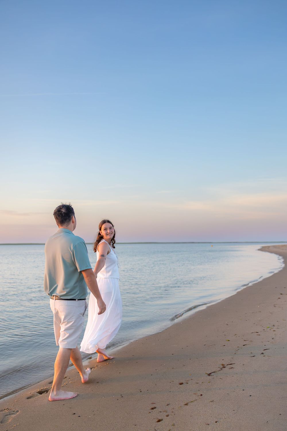Couple walking barefoot on sandy beach at sunset, woman in white dress holding hands with man in casual attire.