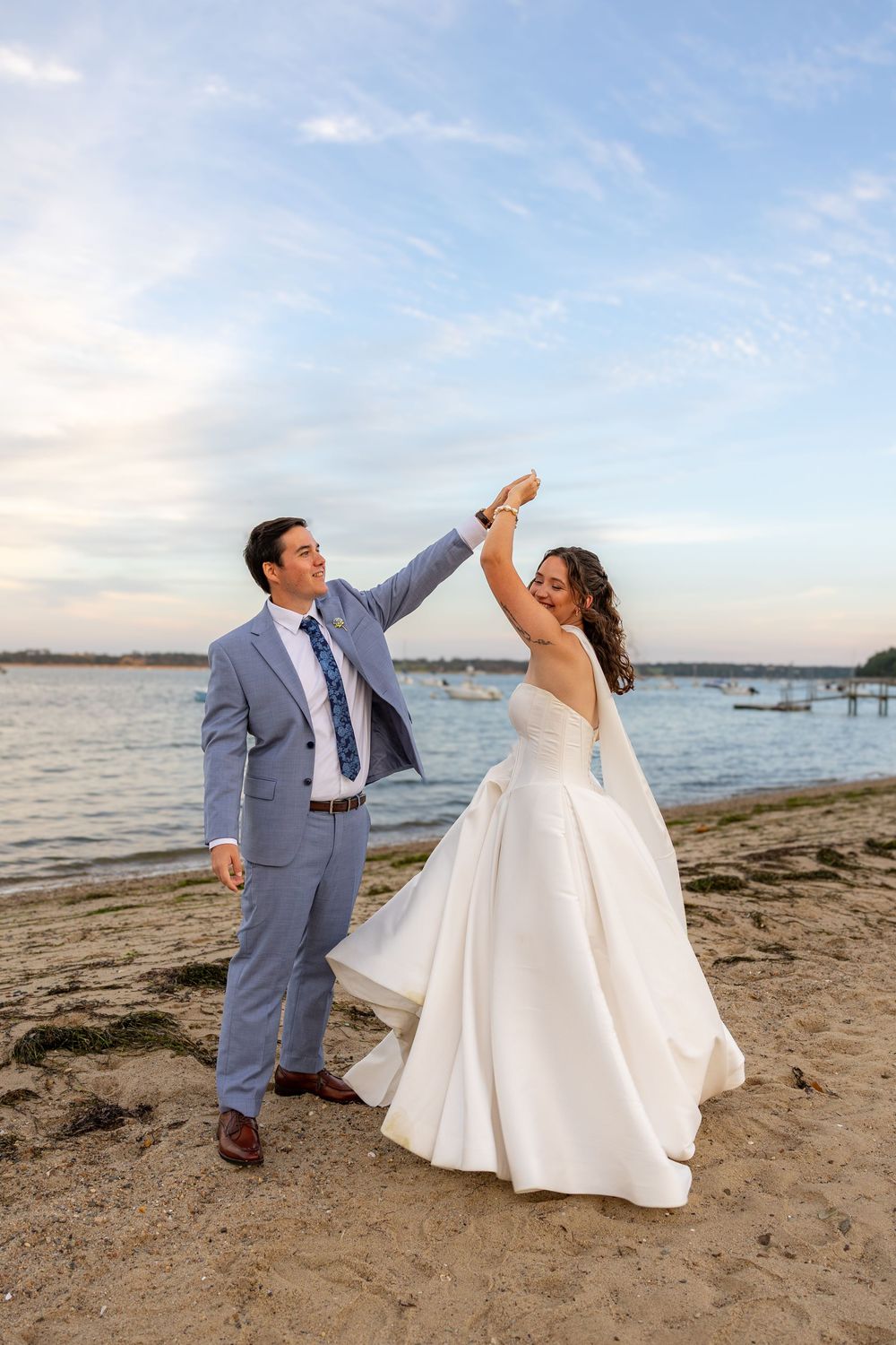 Bride and groom dancing on a sandy beach at sunset, groom in blue suit twirling bride in white gown.