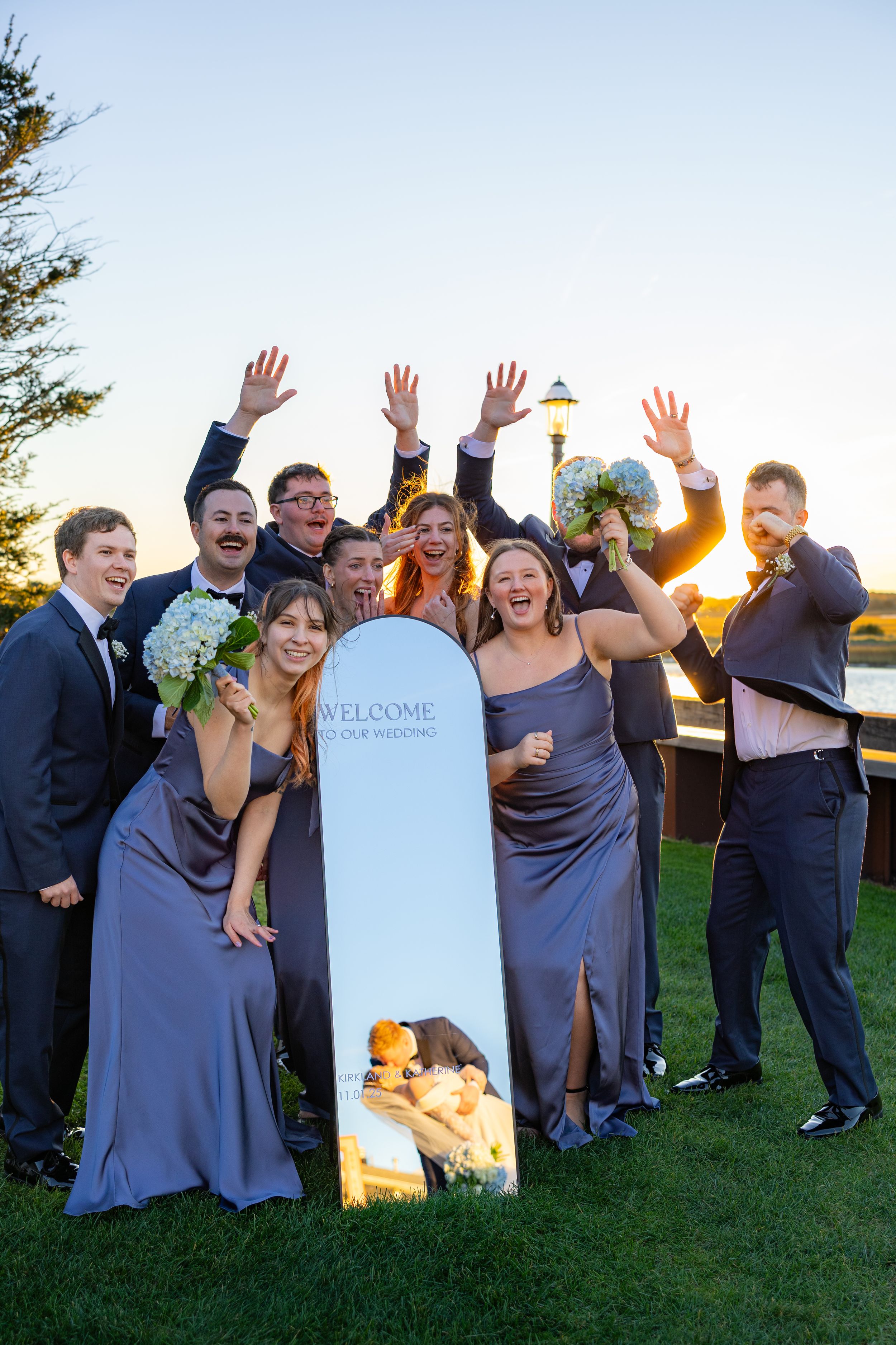Wedding party in purple dresses and dark suits poses playfully by a gravestone at sunset.