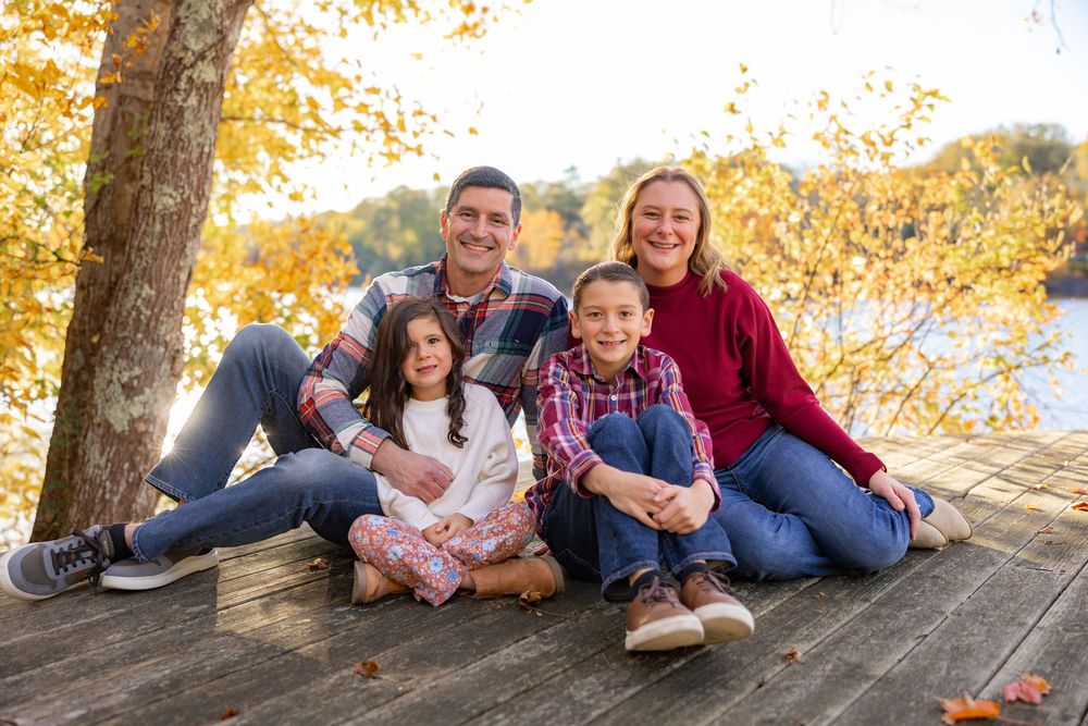 A family in casual attire poses together on a wooden deck surrounded by autumn foliage on a sunny day.