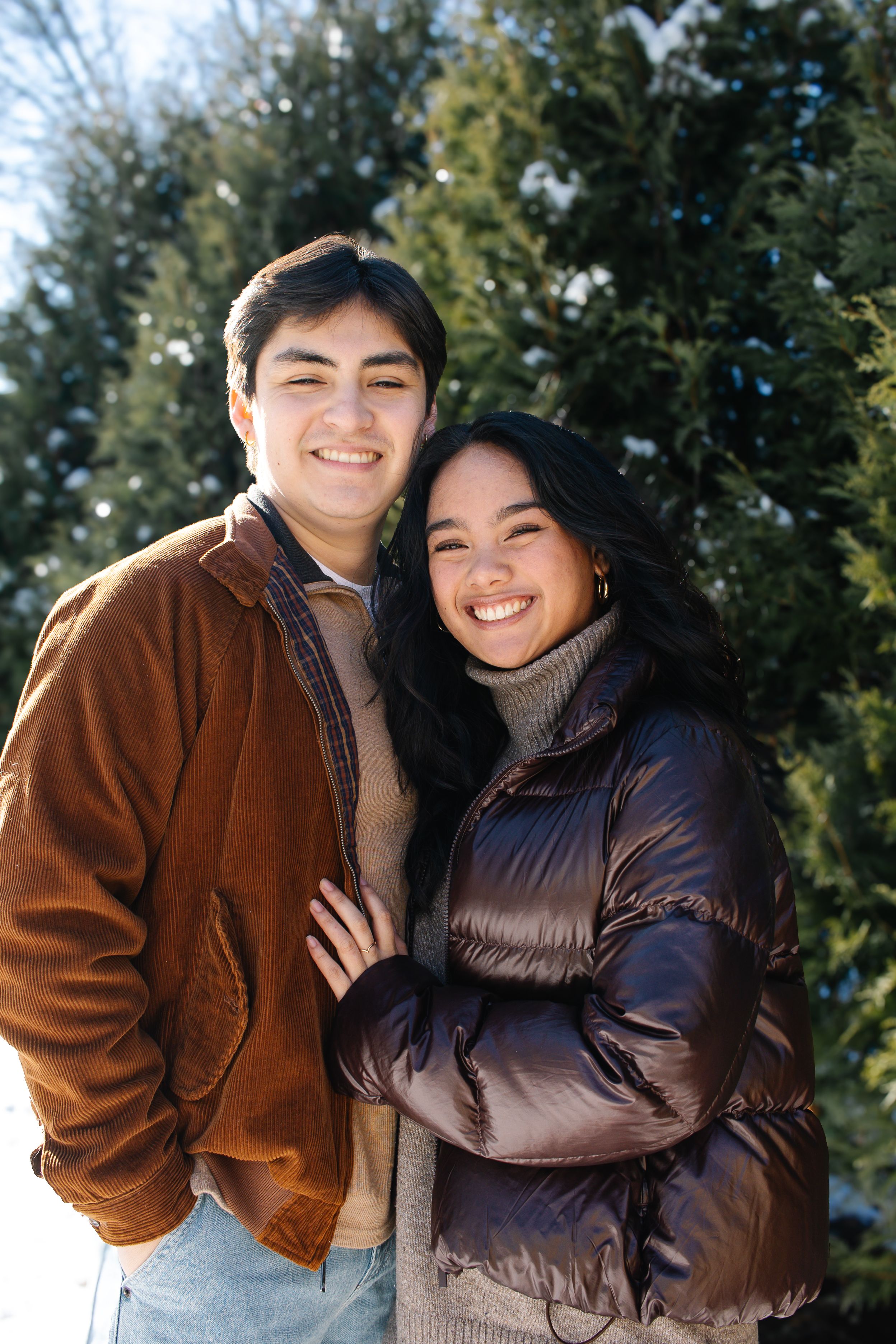 A couple in winter coats share a joyful embrace against a snowy evergreen backdrop.