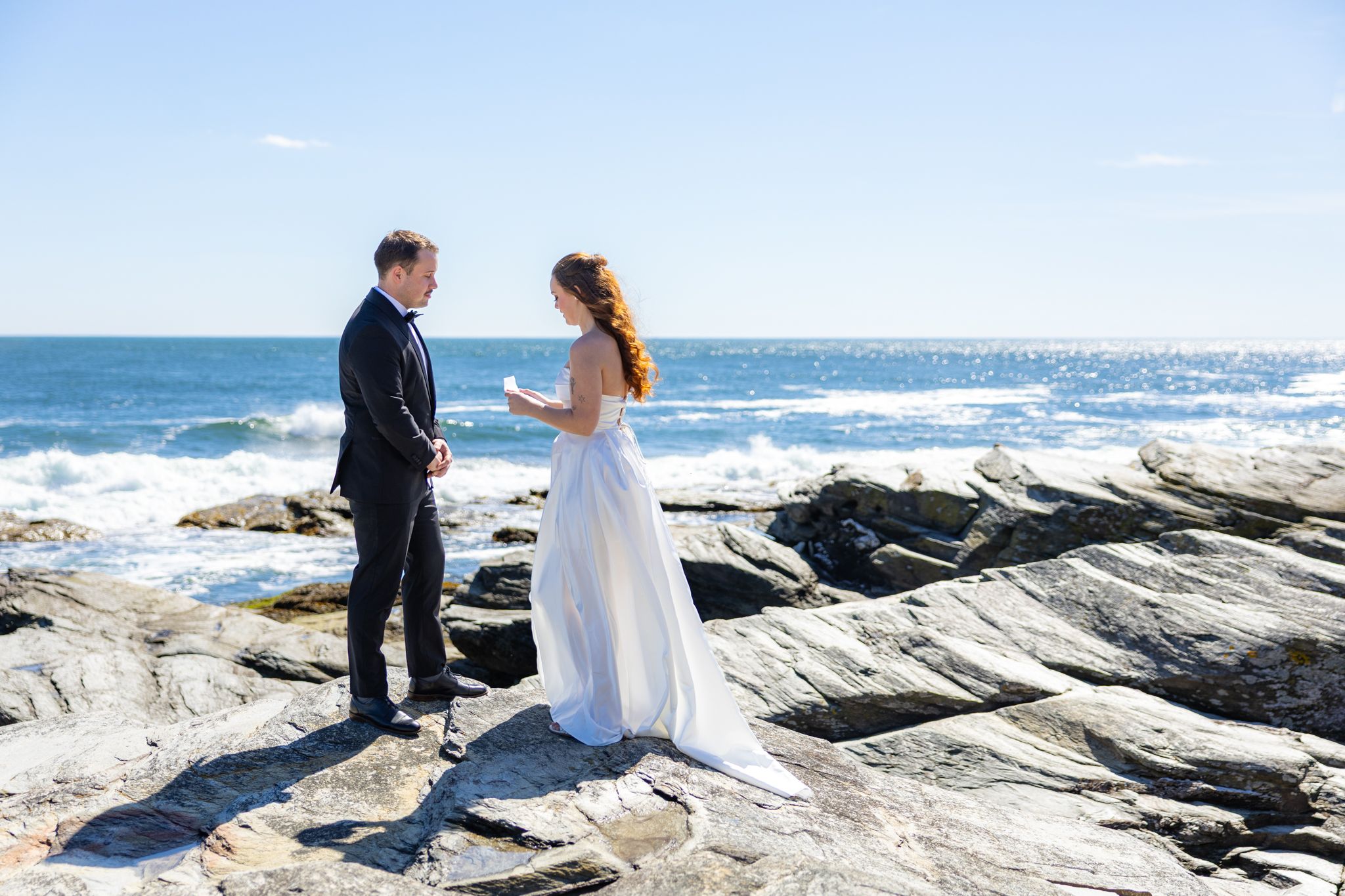 A couple in wedding attire stands together on scenic coastal rocks with ocean waves crashing in the background.