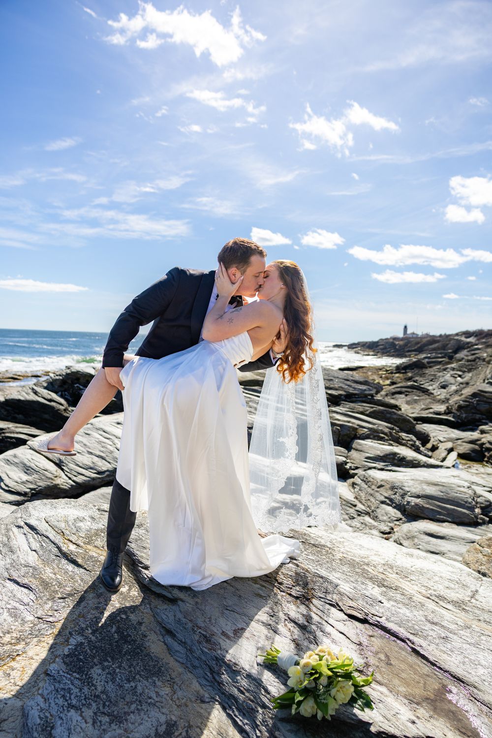 A romantic couple embraces on coastal rocks during a seaside wedding photoshoot with dramatic ocean views and blue skies.