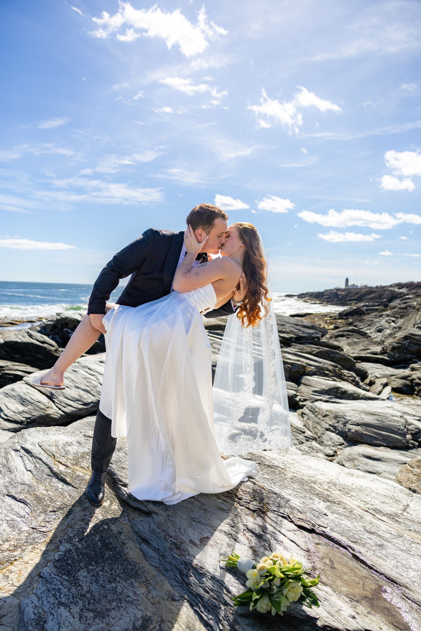 A romantic couple embraces on coastal rocks during a seaside wedding photoshoot with dramatic ocean views and blue skies.