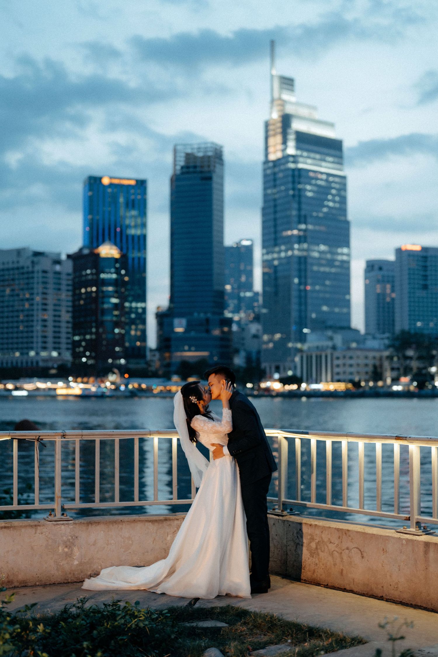 Romantic couple kissing in wedding attire by waterfront with illuminated city skyline at dusk.