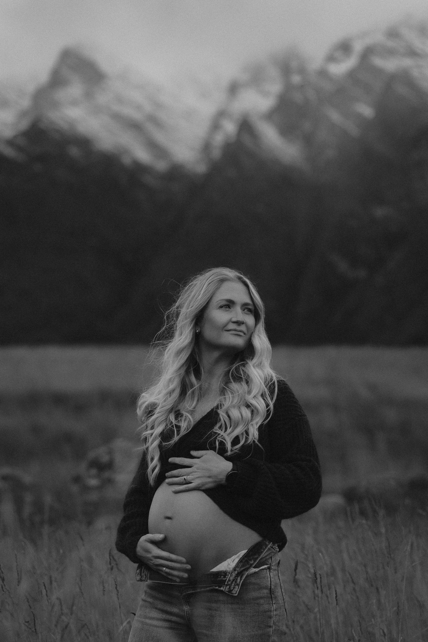 Black and white maternity photo of pregnant woman holding belly in mountain meadow with snow-capped peaks behind her.