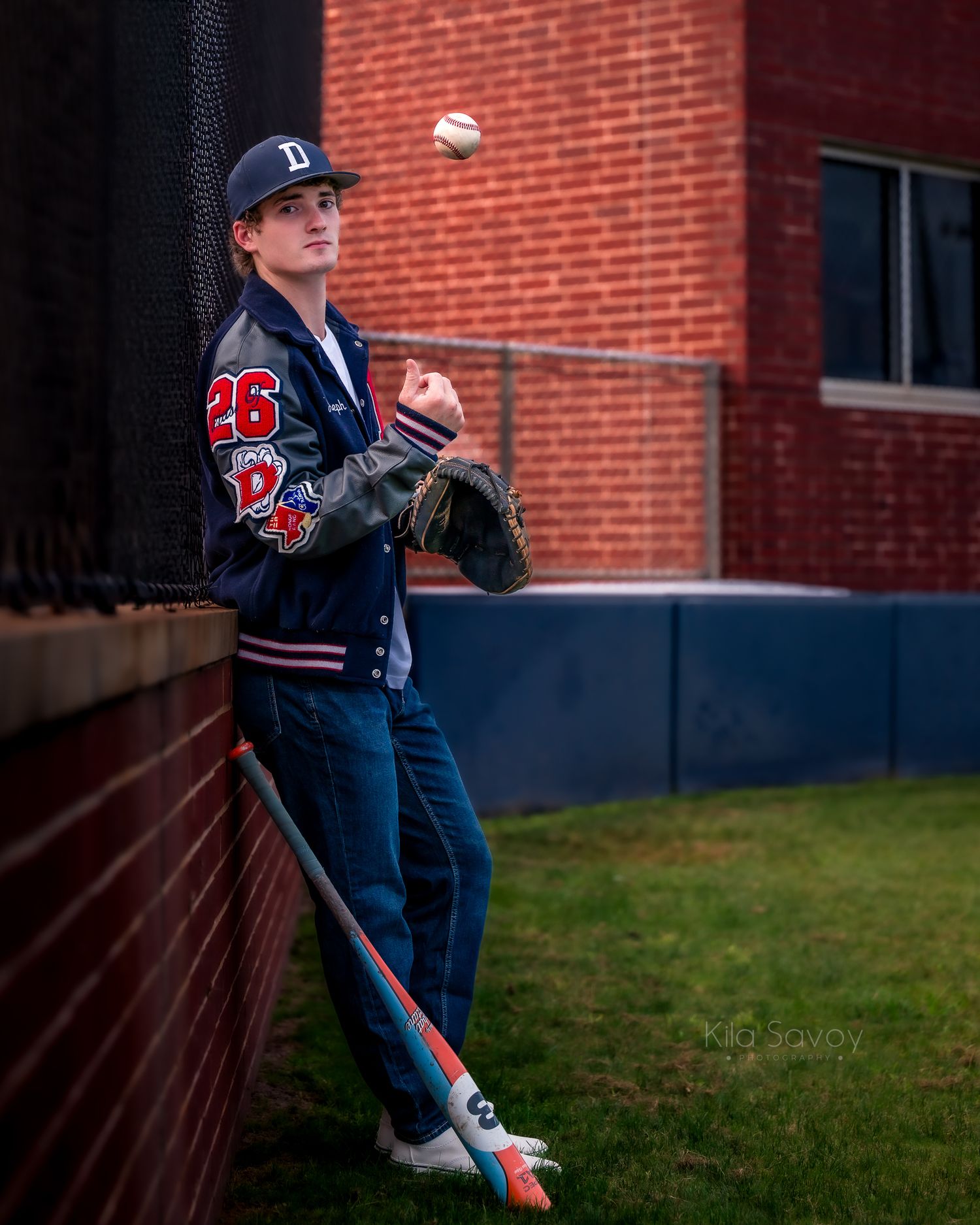 Teen baseball player in navy varsity jacket tossing a ball, holding a bat and glove near a brick wall.