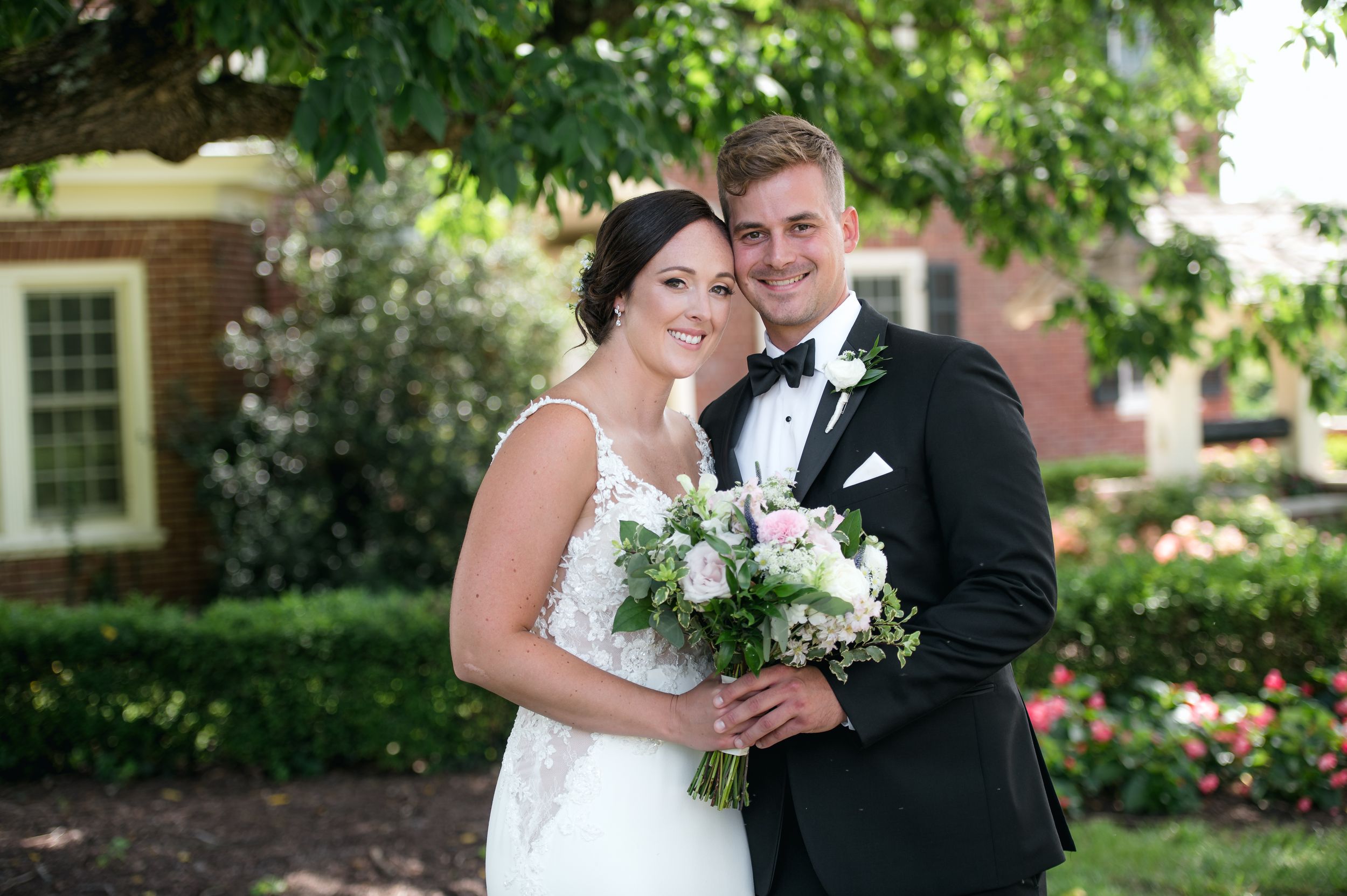 Smiling bride in lace gown holds pink and white bouquet beside groom in black tuxedo outdoors.