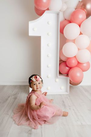 One-year-old baby girl in a pink tulle dress sitting beside a marquee number one with pink balloons.