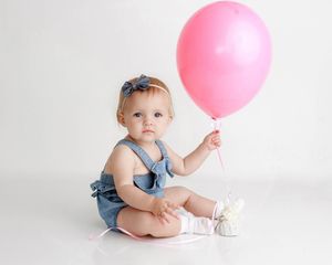 Adorable baby girl in denim romper holding a pink balloon while sitting on a white background.