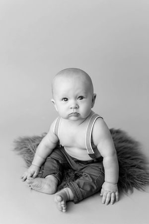 Black and white portrait of a baby boy sitting up wearing suspenders and pants on a fluffy rug.