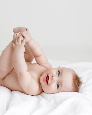 Happy baby lying on white blanket holding feet up smiling during a 6 month old studio portrait session.