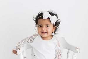 Smiling toddler girl in white lace dress with white bow headband in studio portrait.