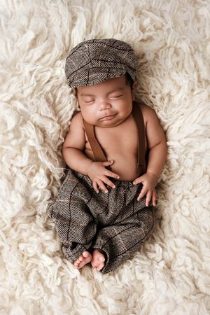 Sleeping newborn baby dressed in tweed newsboy cap and matching pants on fluffy cream rug.