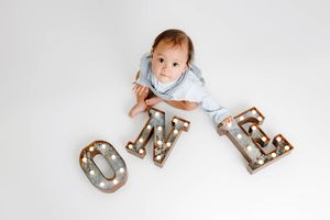 Baby boy in blue onesie looks up at camera while leaning on lit marquee ONE letters.