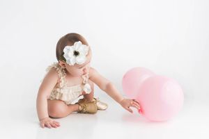 Baby girl in gold outfit and white flower headband reaching for pink balloons during first birthday photo session.